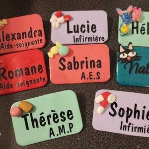 May include: A collection of colorful, handmade name badges. Each badge features a name and profession, such as "Lucie, Infirmière" and "Nathalie, A.M.P.", with decorative embellishments like flowers and candies.