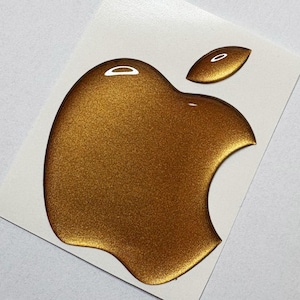 May include: A gold-colored Apple logo sticker, featuring the iconic apple shape with a bite taken out of it and a small leaf. The sticker is on a white rectangular backing.