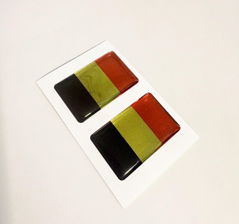 May include: Two rectangular stickers featuring the Belgian flag design. Each sticker has three vertical stripes in black, gold, and red. The stickers are glossy and set on a white background.