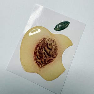 May include: A sticker featuring a peach-themed design. The sticker is shaped like an apple with a bite taken out, revealing a peach pit. A green leaf is positioned above the peach. The sticker has a glossy finish.