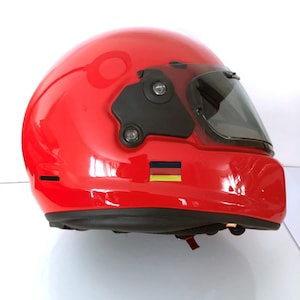 May include: A glossy red motorcycle helmet with a dark visor and black accents. The helmet features a small German flag sticker on the side. The helmet is designed for safety and protection.