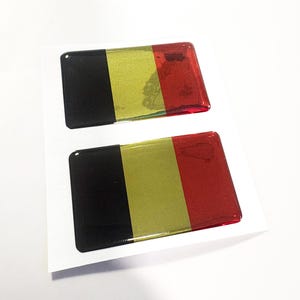 May include: Two rectangular stickers featuring the Belgian flag design. Each sticker has a black, gold, and red vertical stripe pattern. The stickers are on a white background.