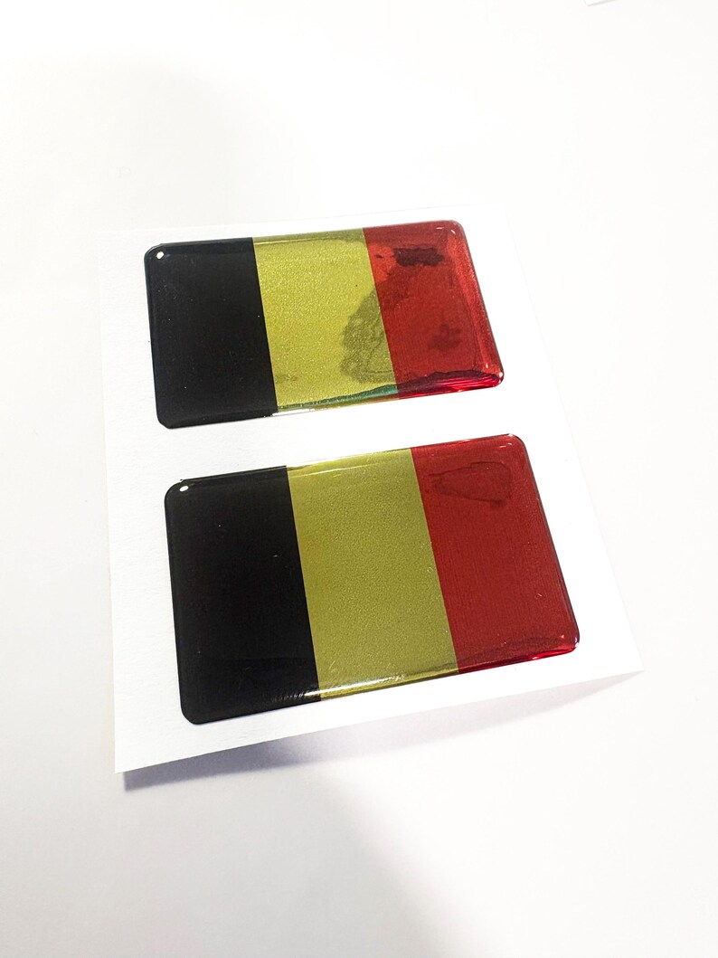 May include: Two rectangular stickers featuring the Belgian flag design. Each sticker has a black, gold, and red vertical stripe pattern. The stickers are on a white background.