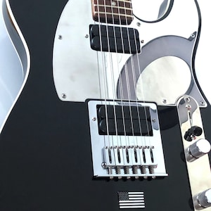 May include: Black electric guitar with a silver pickguard and hardware. The guitar features a small American flag emblem. The body is glossy black, and the neck has a brown fretboard. The guitar has six strings.
