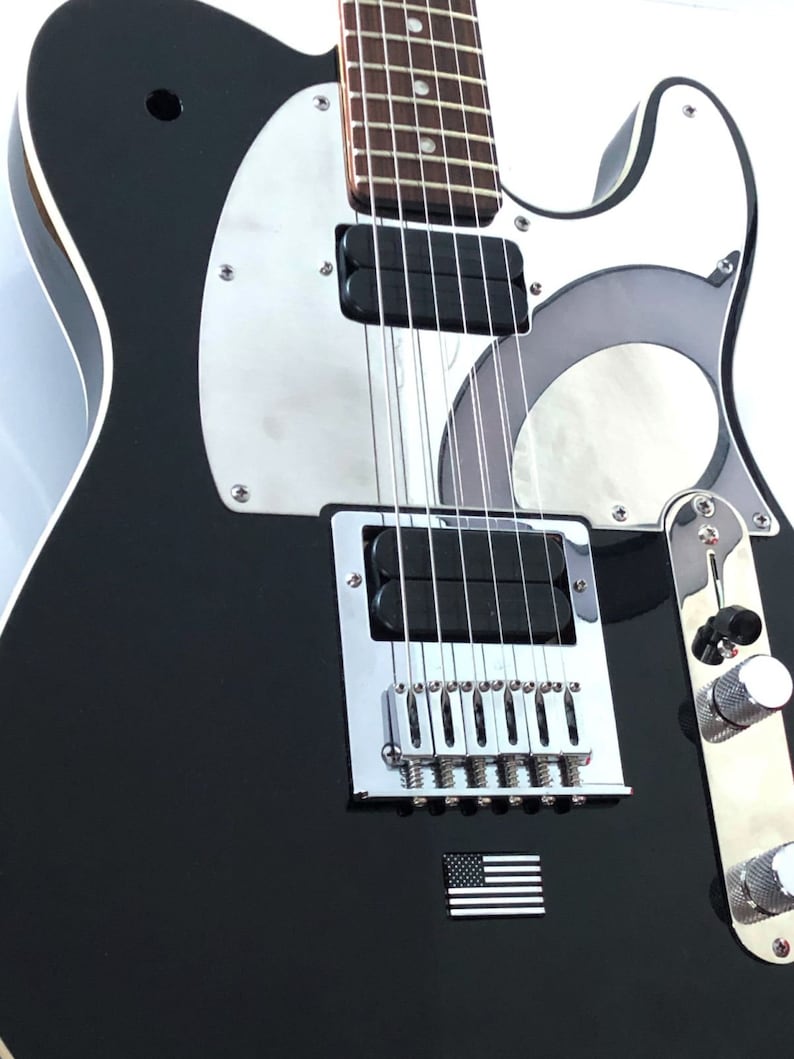 May include: Black electric guitar with a silver pickguard and hardware. The guitar features a small American flag emblem. The body is glossy black, and the neck has a brown fretboard. The guitar has six strings.