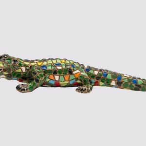 May include: A mosaic alligator sculpture in shades of green, blue, orange, and yellow. The reptile's body is covered in colourful tiles, with an open mouth revealing teeth. The sculpture is a decorative item.