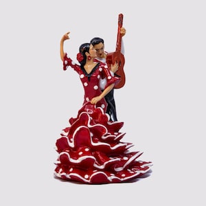 May include: A figurine of a flamenco dancer in a red dress with white polka dots, and a man playing a guitar. The woman has her arm raised, and the man is holding a brown guitar. The figurine is a souvenir.