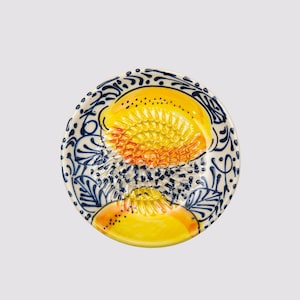 Puede incluir: Un rallador de cerámica con diseño de limón. El rallador presenta un diseño de limón amarillo y naranja en el centro, rodeado de un patrón floral azul y blanco. El rallador es redondo y tiene una superficie texturizada.
