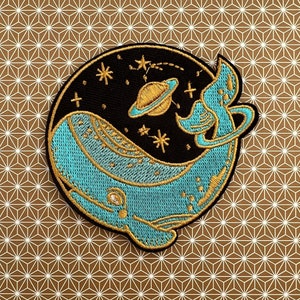 Könnte beinhalten: Ein gestickter Aufnäher mit einem blauen Wal, der im Weltraum mit einem Planeten und Sternen schwimmt, in goldener und blauer Farbe.