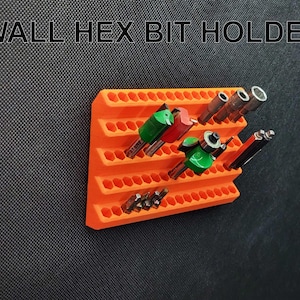 Op de afbeelding: Een oranje wandhouder voor zeskantbits, gevuld met diverse boorbits. De houder heeft meerdere rijen zeshoekige gaten om verschillende bitmaten te herbergen. De tekst "WALL HEX BIT HOLDER" staat bovenaan.