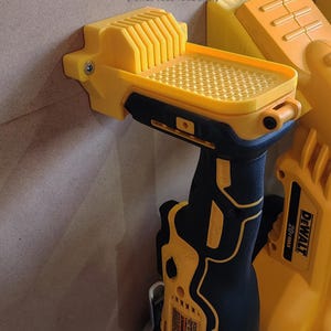 Peut inclure: Support mural jaune pour outil oscillant de 20 volts. Le support a une surface texturée et une fente pour la lame de l'outil. Le texte "OSCILLATING TOOL HOLDER (FOR 20 VOLS TOOL ONLY)" est imprimé sur le mur au-dessus du support. Le texte "(TOOL NOT INCLUDED)" est imprimé sous le support.