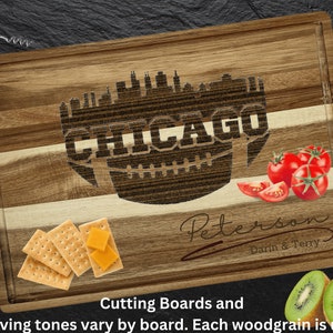 Puede incluir: Tabla de cortar de madera con el horizonte de Chicago grabado en el centro. La palabra "CHICAGO" está grabada en forma de balón de fútbol. Incluye tomates, galletas, queso y kiwi. Un sacacorchos está en la esquina superior izquierda.