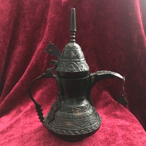 May include: A black metal coffee pot with intricate carvings and a decorative lid. The pot has a spout and a handle.