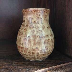 May include: A small, handcrafted ceramic vase with a textured surface. The vase features a speckled pattern in shades of tan and brown, with a slightly flared rim. The vase is displayed on a wooden shelf.