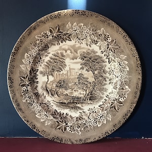 May include: A brown and white ceramic plate with a floral design and a depiction of a castle in the centre. The plate features a detailed design of leaves and flowers.