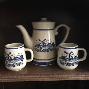May include: A set of three ceramic teacups and a teapot with a blue and white windmill design. The teapot has a handle and a spout. The teacups have handles. The design features a windmill, houses, and a river.