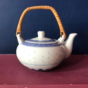 May include: A white teapot with a blue and white floral pattern and a woven bamboo handle. The teapot has a round body and a spout on the side.