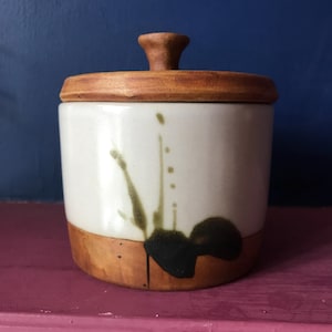 May include: A white ceramic jar with a wooden lid and brown and green abstract design. The jar is sitting on a pink surface.