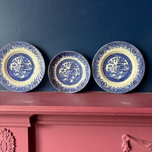 May include: Three blue and white ceramic plates featuring the Willow pattern. Each plate displays a detailed landscape scene within a circular border. The plates are arranged in a row against a dark blue backdrop, typical of traditional tableware.