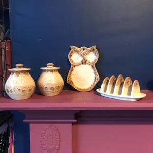 May include: A set of four ceramic kitchenware items in a brown and white glaze. The set includes two lidded jars, an owl-shaped dish, and a toast rack.