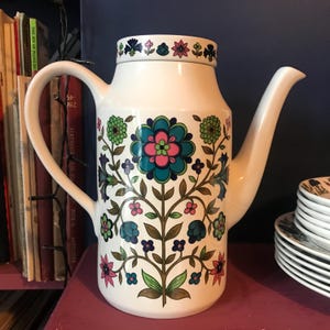 May include: White ceramic coffee pot with a floral design in shades of green, blue, pink, and brown. The pot has a lid and a spout.
