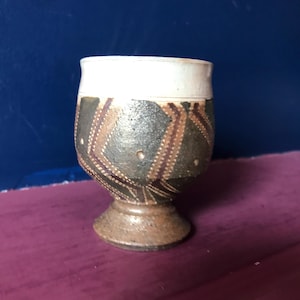 May include: A small, brown and white ceramic goblet with a geometric pattern. The goblet has a wide base and a narrow top.