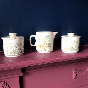 May include: A white ceramic sugar bowl and creamer set with a floral pattern in shades of green and gold. The set has a lid and a spout.