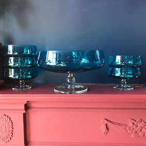 May include: A selection of teal glass serving bowls and dishes. The set includes a large centerpiece bowl and several smaller bowls, all with a pedestal base. The bowls are arranged on a pink surface against a dark blue background.