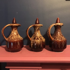 May include: Three brown and cream ceramic jugs with a mottled glaze. The jugs have a rounded body and a spout on one side. The jugs are arranged in a row on a pink shelf.
