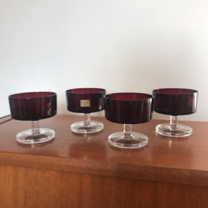 May include: Four vintage dessert glasses with stems. Each glass has a clear, circular base, a thin stem, and a wide, cylindrical bowl in a deep burgundy colour. The glasses are displayed on a wooden surface against a white backdrop.