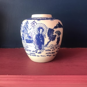 May include: A small, cream-coloured ceramic vase with a blue and white Asian-inspired design. The vase features a detailed illustration of figures with umbrellas, trees, and mountains. The design wraps around the vase.