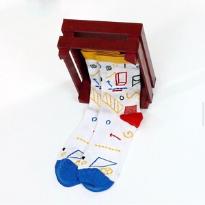 May include: A pair of white socks with a colourful geometric pattern, including red, yellow, blue, and black. The socks are folded and placed in a red wooden crate.