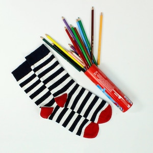 Op de afbeelding: Een paar zwart-wit gestreepte sokken met rode harten op de tenen. De sokken liggen op een wit oppervlak naast een rood-witte buis met kleurpotloden.