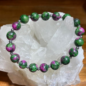 May include: A beaded bracelet featuring round gemstones with a mix of green and magenta colors. The beads are strung together with small silver-colored spacers. The bracelet is displayed on a white, crystalline surface.