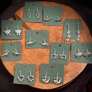 Puede incluir: Una colección de pendientes colgantes plateados exhibidos en tarjetas verdes, dispuestos sobre una superficie de madera. Los pendientes presentan varios diseños inspirados en animales y la naturaleza, incluyendo peces, ovejas, dinosaurios, elefantes, tortugas, delfines, perezosos y abejas.