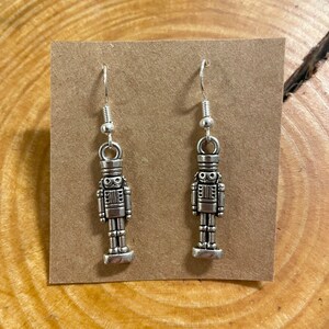 Puede incluir: Un par de pendientes colgantes de metal plateado. Cada pendiente presenta un diseño detallado de soldado cascanueces en miniatura. Los pendientes se exhiben en una tarjeta marrón.
