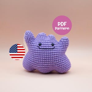 May include: A handmade, purple crochet plush toy with a smiling face and a unique shape. The toy has two small black eyes and a stitched mouth. A pink circle with the text "PDF Pattern" is in the background. An American flag is also in the image.