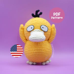 Puede incluir: Un peluche de Psyduck amarillo tejido a mano con ojos blancos, un pico blanco y un mechón de pelo negro. El peluche está sobre un fondo morado. La imagen también incluye una bandera estadounidense circular y un círculo rosa con el texto "PDF Pattern".