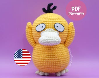 May include: A handmade, yellow crochet Psyduck plushie with white eyes, a white beak, and a black tuft of hair. The plushie is standing on a purple background. The image also includes a circular American flag and a pink circle with the text "PDF Pattern".