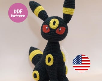May include: Crocheted Umbreon plushie in black with yellow accents, red eyes, and a round yellow design on its body. The plushie has long ears and a tail with yellow stripes. The image includes a pink circle with "PDF Pattern" text and a US flag.