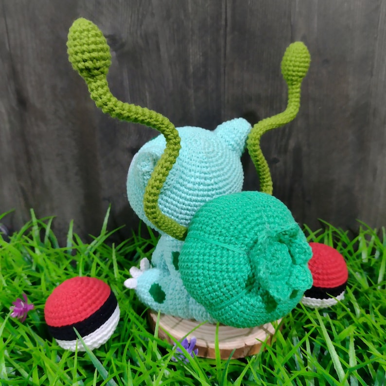May include: A crocheted light blue creature with green tendrils and a green ball, on a wooden disc. Two red and white crocheted balls are on the grass. This handmade item is a unique, whimsical creation.