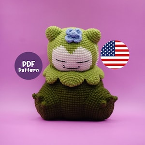 Puede incluir: Un peluche verde hecho a mano con ganchillo, con una cara blanca, ojos cerrados y una pequeña flor azul en la cabeza. El juguete está sentado y tiene forma redonda. La imagen también incluye una etiqueta de patrón PDF y una bandera estadounidense.