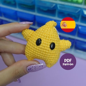 Puede incluir: Una estrella amarilla de ganchillo hecha a mano con ojos de botón negros. La estrella tiene un diseño de cinco puntas. La imagen incluye una bandera española y un círculo morado con el texto "PDF Patrón".