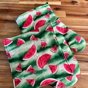 May include: A green and white oven mitt and pot holder set with a watermelon slice pattern. The design features red watermelon slices on a green background. The oven mitt has a loop for hanging.