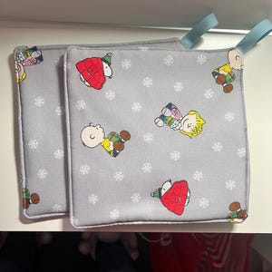 May include: Two gray pot holders with a white snowflake pattern and a repeating image of Charlie Brown and Snoopy in winter clothing.