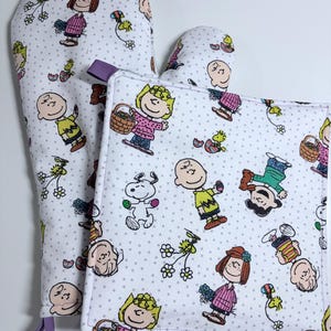 May include: White oven mitt and pot holder set featuring a repeating pattern of cartoon characters, flowers, and Easter eggs on a dotted background. The oven mitt has a purple loop for hanging.