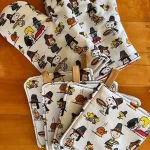 May include: Set of six white oven mitts with a Thanksgiving theme featuring Peanuts characters dressed as pilgrims. The mitts have a brown trim and are decorated with images of Snoopy, Charlie Brown, Lucy, and other Peanuts characters.