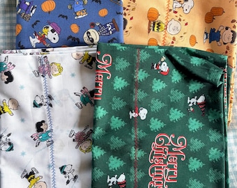 May include: Four folded fabric panels featuring Peanuts characters. One blue with pumpkins and bats, one orange with pumpkins and leaves, one white with winter skating scenes, and one green with Christmas trees and the text Merry Christmas.