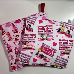 May include: Two Valentine's Day themed pot holders. One is pink with gnome illustrations and the other is white with pink hearts and Peanuts comic strip quotes. Both have a loop for hanging.