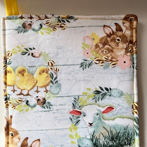 May include: A yellow and white kitchen pot holder with a farmhouse theme. The pot holder features a white background with a woodgrain pattern and images of bunnies, chicks, and a lamb. The pot holder has a yellow border.